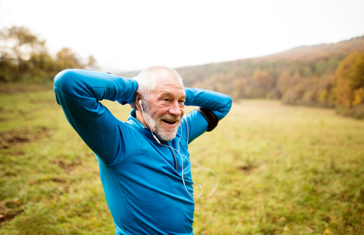 Senior runner in sunny autumn nature doing stretching. Man with earphones, listening music, armband on his arm. Rear view.